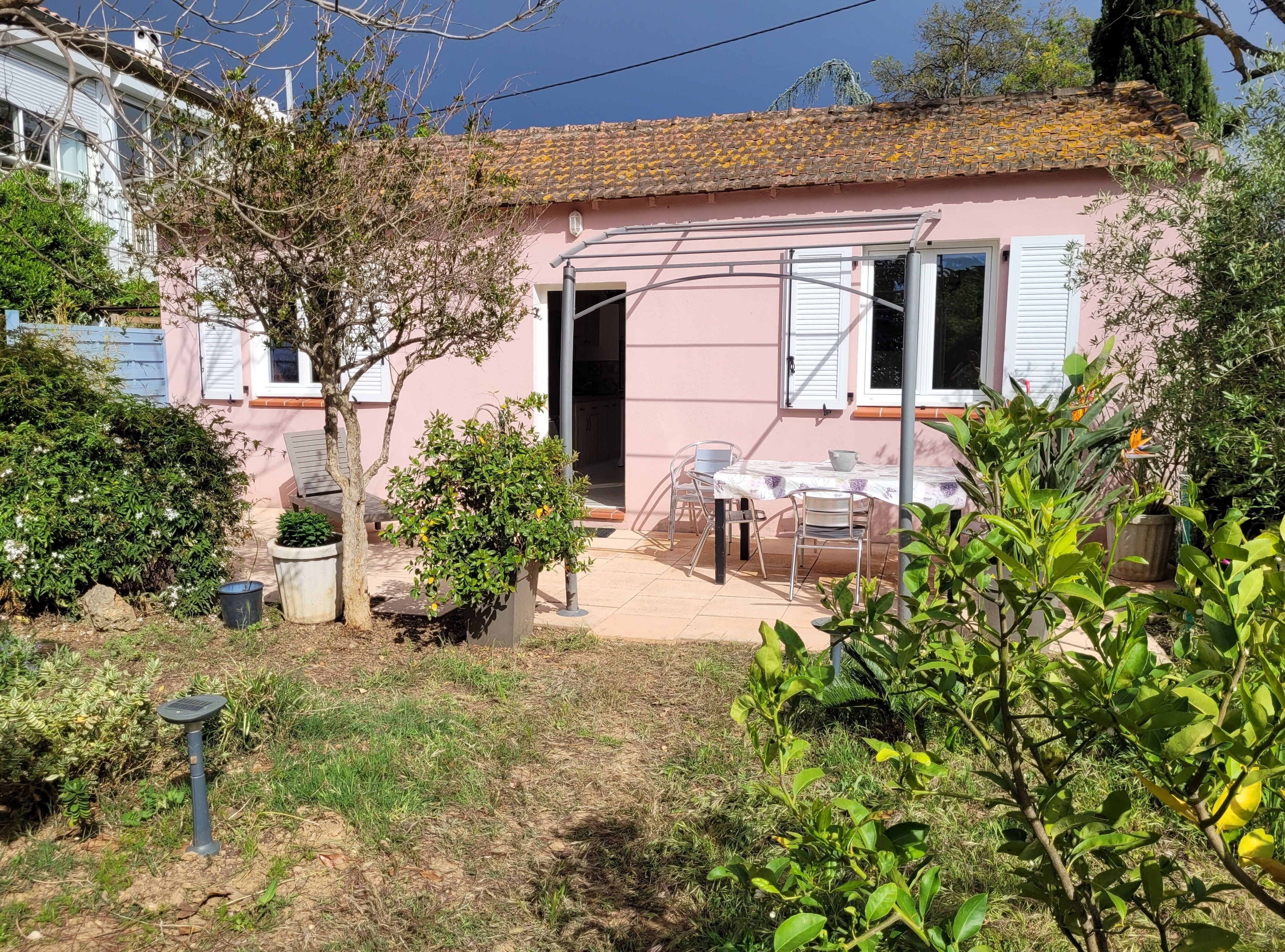 A pink house with a tiled roof, a patio with a table and chairs, and various potted plants.