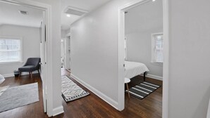 Une pièce spacieuse et bien éclairée, dotée d’un plancher en bois, d’une grande fenêtre avec des stores, d’un lit recouvert d’une courtepointe blanche, d’une chaise et d’un tapis à motifs.