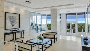 A spacious, modern lobby with a glass wall offering a view of the outdoors, a seating area with a glass coffee table, and a framed artwork on the wall. - Beachfront Condo With Views, Pools and Tennis Courts (Marco Island)