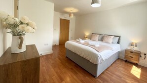 A bedroom with a wooden floor, a bed with white bedding, a bedside table with a lamp, and a vase with flowers on a wooden dresser.
