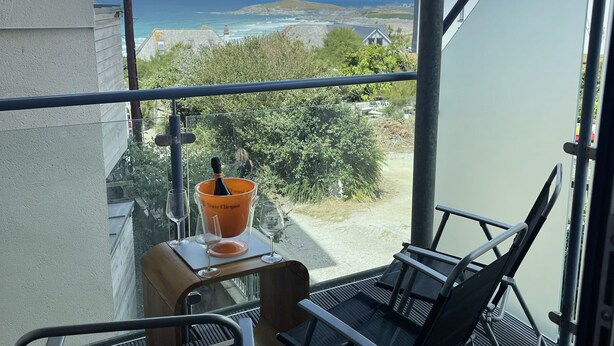 A balcony with a view of the ocean, a table with a bucket and wine glasses, and a chair.