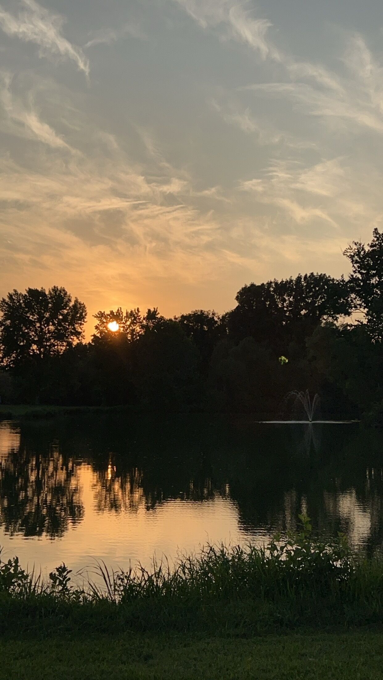 A sunset over a calm lake with trees in the background and a fountain in the water.