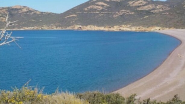 Une route côtière menant à une plage aux eaux d’un bleu limpide, avec des montagnes qui se dessinent à l’horizon.