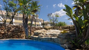A swimming pool surrounded by stone walls and tropical plants, with a clear blue sky in the background.