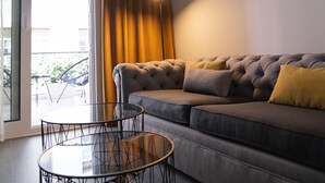 A modern living room with a grey sofa, a round glass table, and a view of the outdoors through a sliding glass door. - K23 Color Suites (Athens)