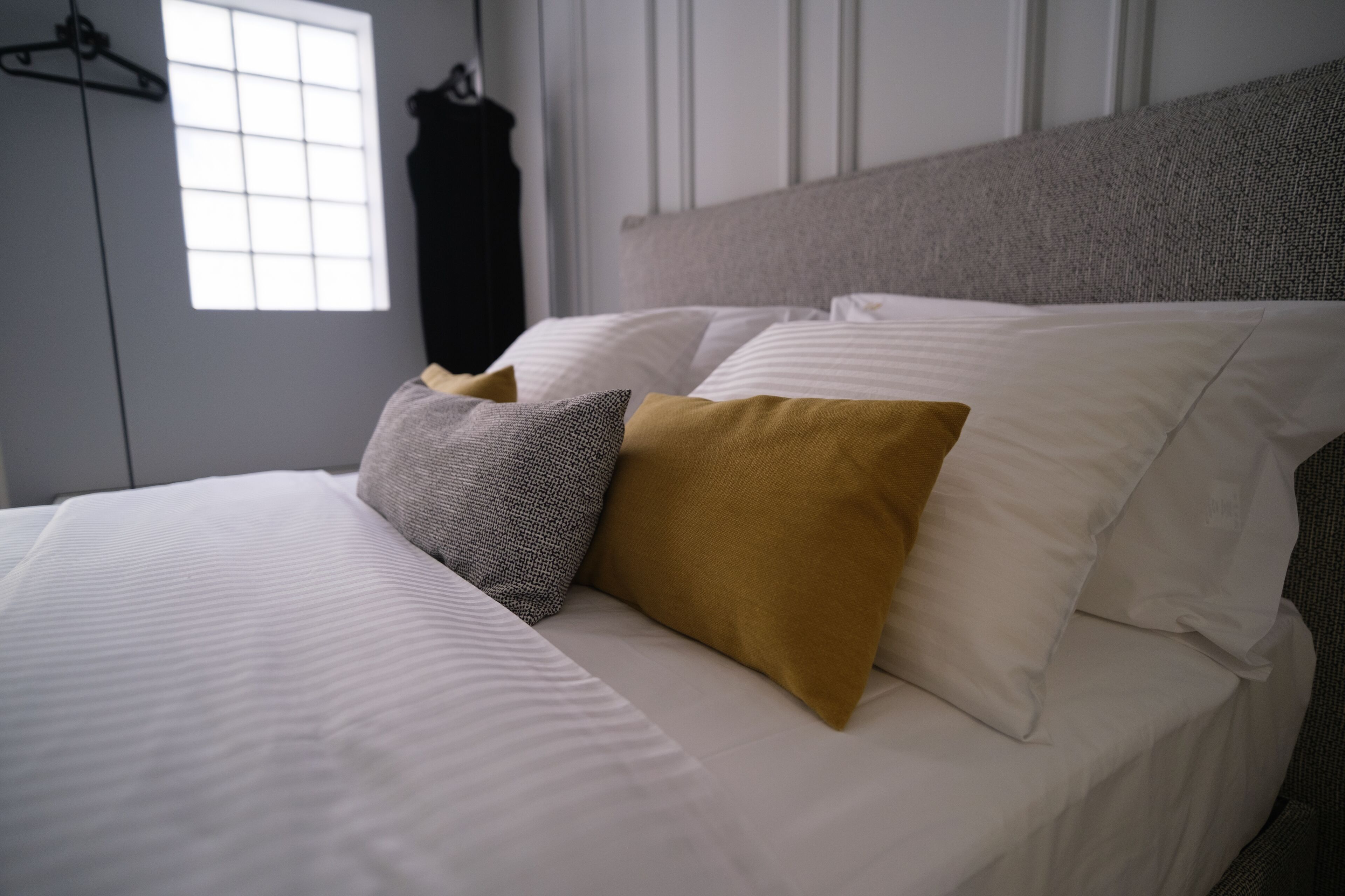 A neatly made bed with white and mustard-colored pillows, a grey pillow, and a grey headboard.