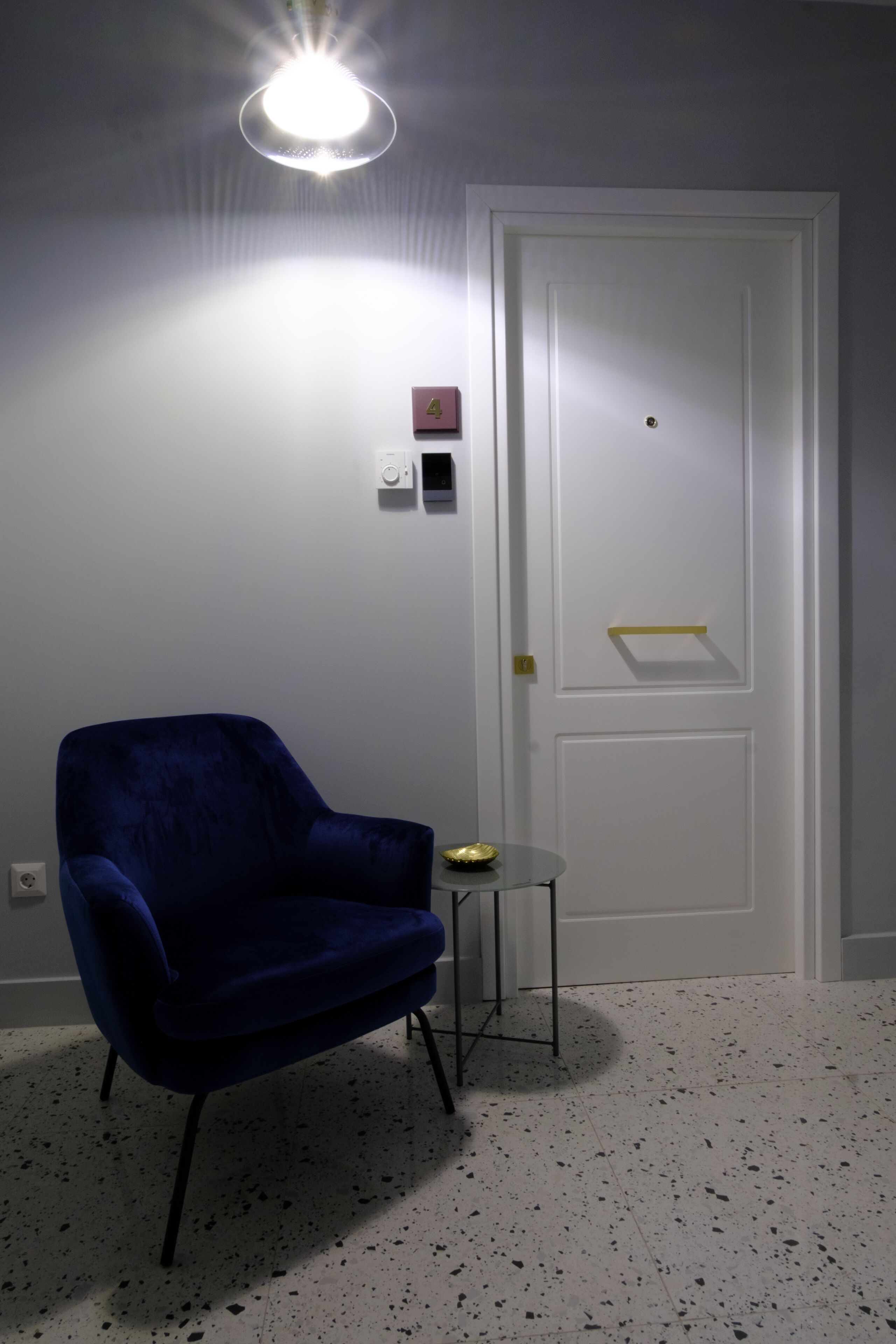 A room with a blue chair, a small table, a white door, and a light fixture on the ceiling.