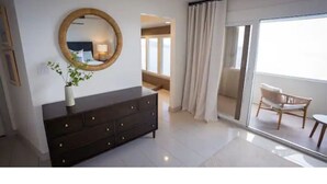 Ein modernes Schlafzimmer mit einer dunklen Kommode, einem runden Spiegel und einem Blumentopf. Eine Glastür führt auf einen Balkon mit einem Stuhl.
