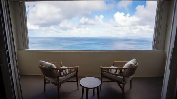 Ein Balkon mit zwei Korbstühlen und einem kleinen Tisch, der auf ein Gewässer und einen bewölkten Himmel blickt.