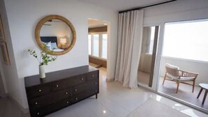 A modern bedroom with a dark dresser, a round mirror, and a potted plant. There is a sliding glass door leading to a balcony with a chair.