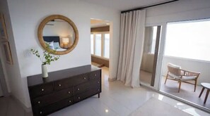A modern bedroom with a dark dresser, a round mirror, and a potted plant. There is a sliding glass door leading to a balcony with a chair.