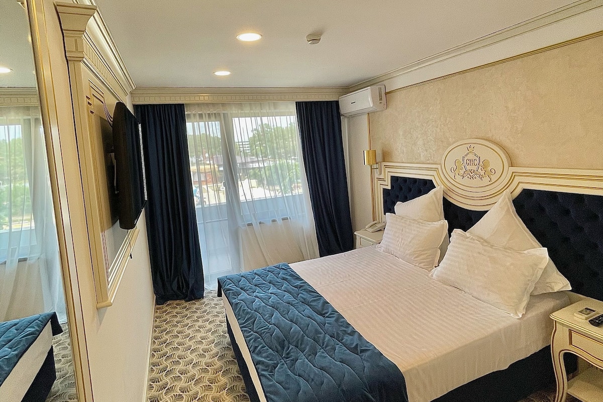 A hotel room with a large bed, a mirror, a bedside table, and a window with curtains.