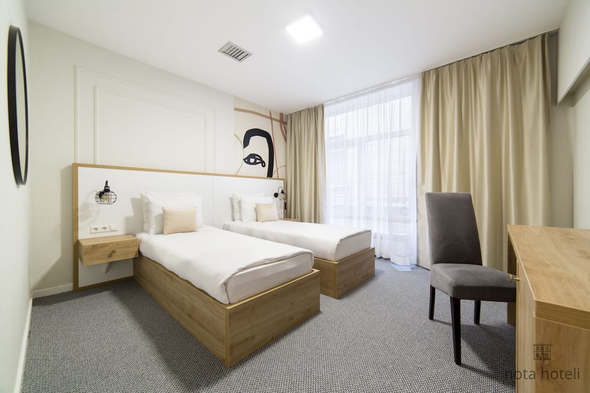 Ett hotellrum med två sängar, ett skrivbord, en stol och ett stort fönster med gardiner.