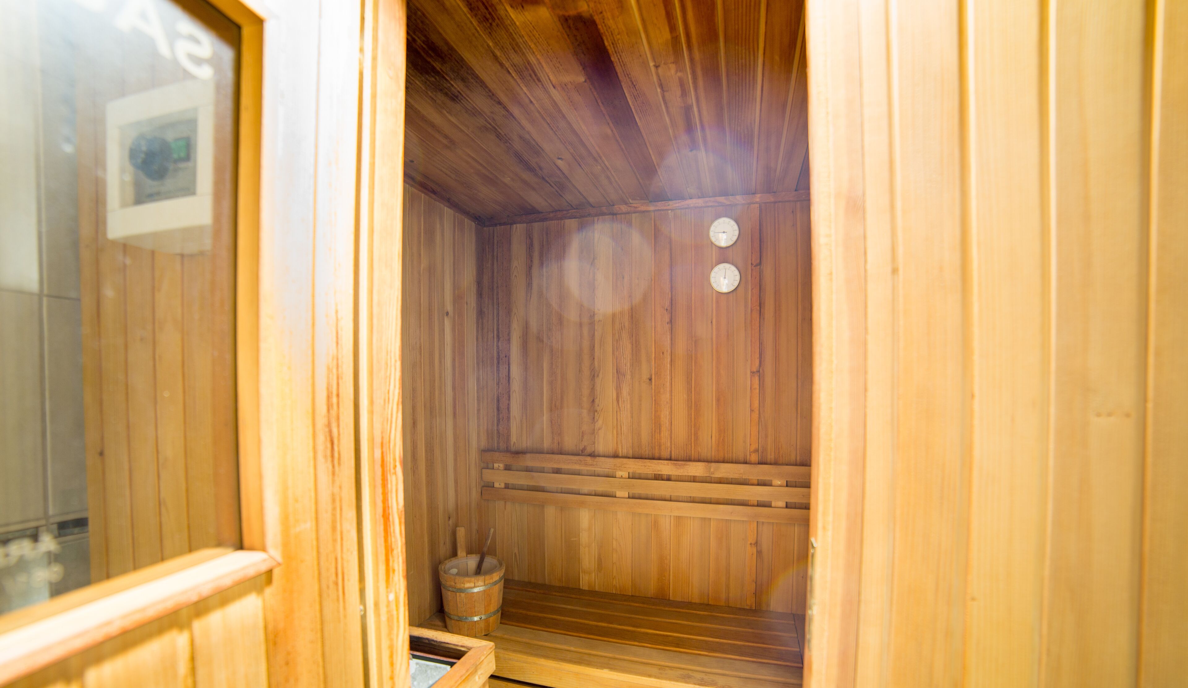 A wooden sauna with a bench, a bucket, and two clocks on the wall.