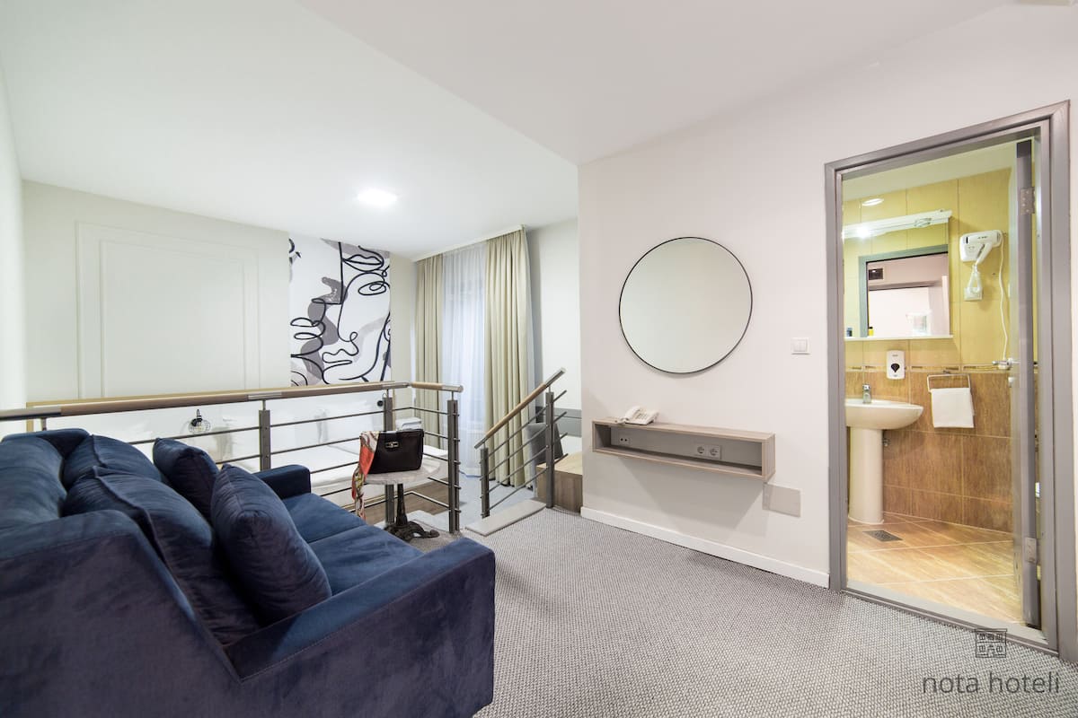 Ett modernt hotellrum med en blå soffa, en trappa, en rund spegel och ett badrum som syns genom en öppen dörr.