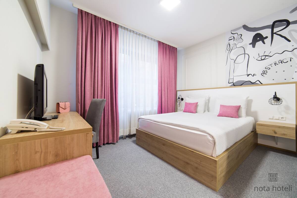 Ett modernt hotellrum med en säng i trä, ett skrivbord med en telefon, en stol och ett stort fönster med rosa gardiner.
