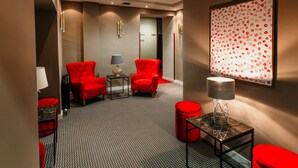 A hotel lobby with a patterned carpet, red upholstered chairs, a table with a lamp, and a framed artwork on the wall. - HOTEL SAHAT (Sarajevo)