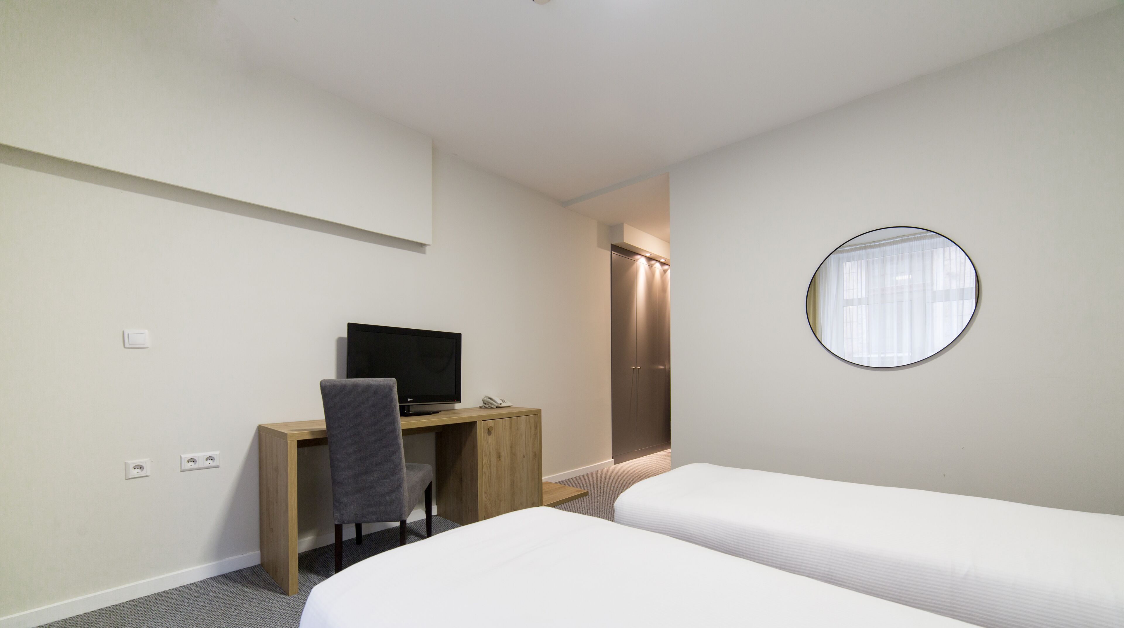 Habitación de hotel con cama, escritorio con computadora, silla, armario y espejo redondo en la pared.