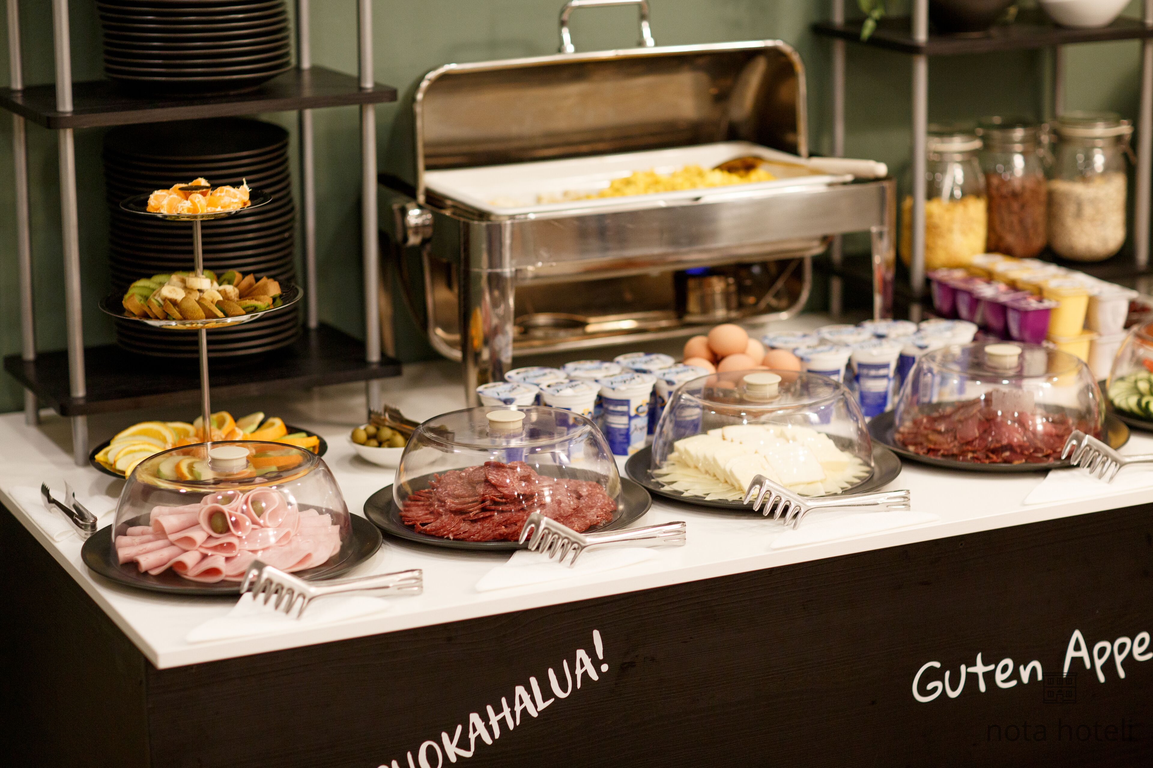 A buffet with various food items and utensils, including plates of sliced meats, cheeses, and fruits.