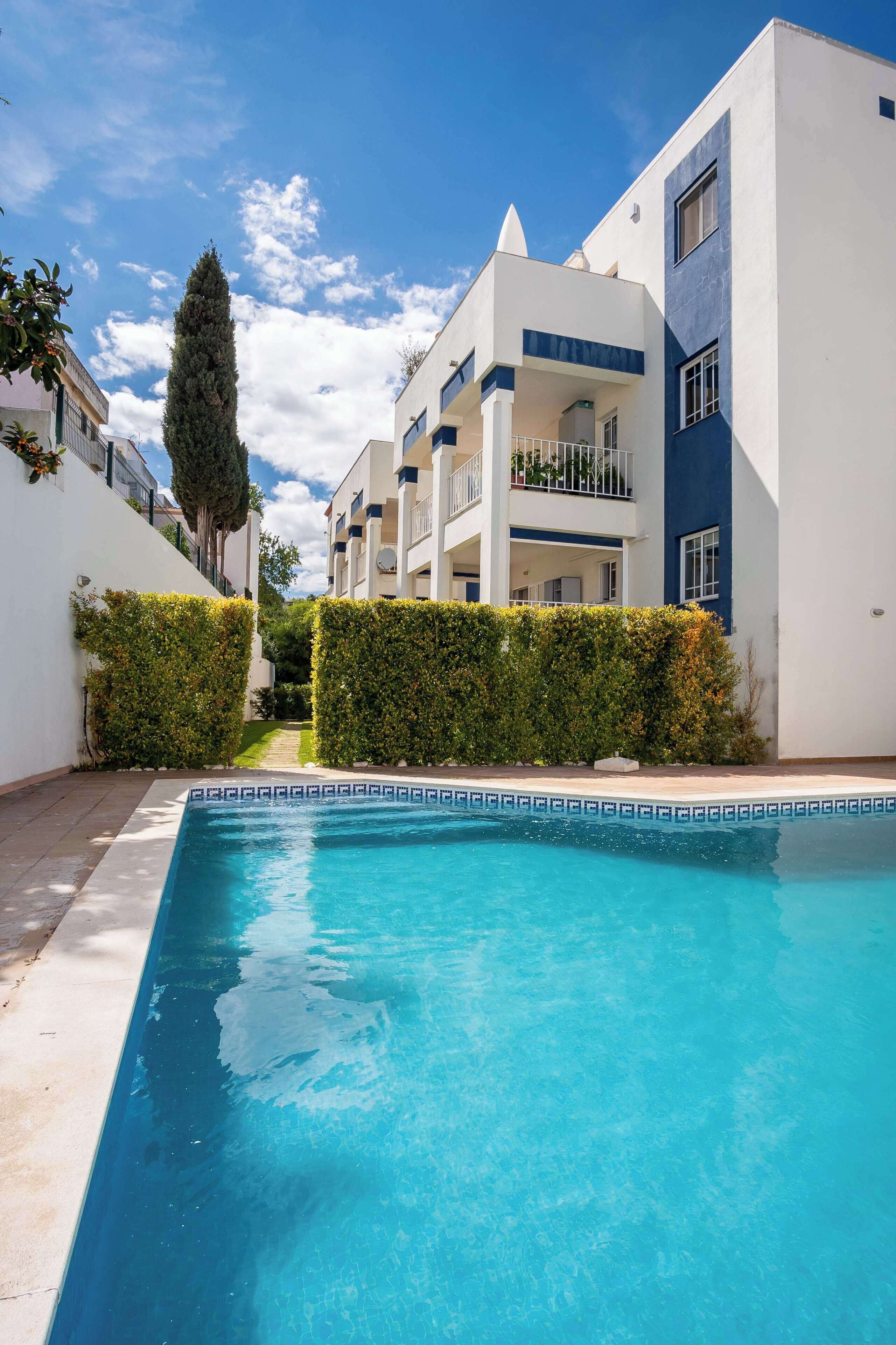 Una piscina con borde de mosaicos geométricos, rodeada de setos cuidados y edificios modernos con balcones.