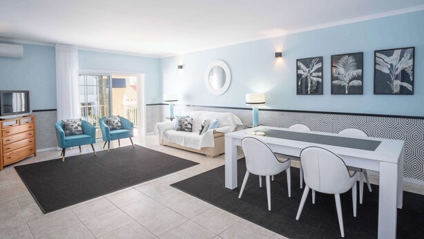 A modern living room with a white sofa, blue armchairs, a dining table with white chairs, and a TV on a wooden cabinet. - Apartment Claro Azul (Albufeira)