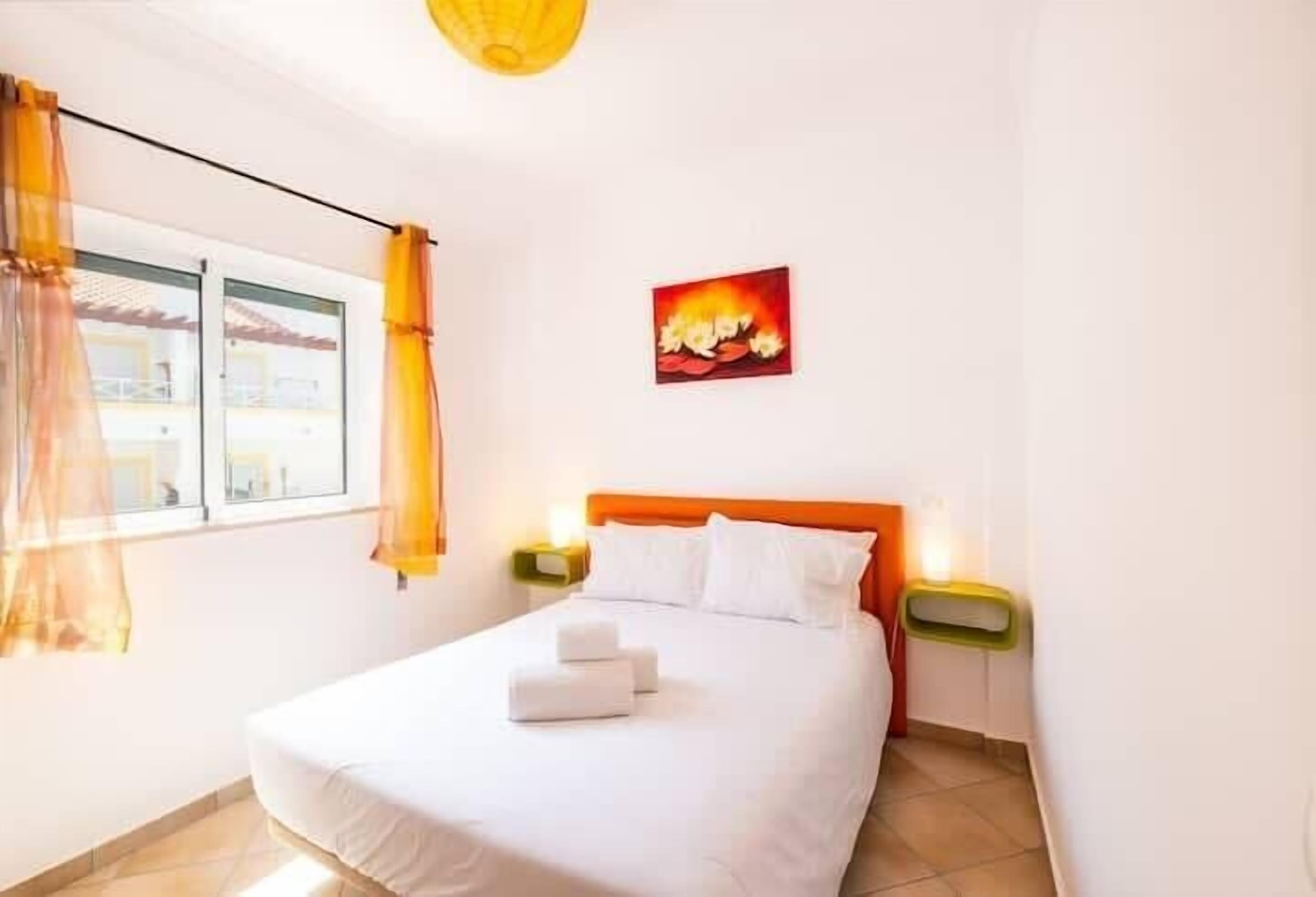 A hotel room with a bed, white curtains, a painting, and a nightstand with a lamp.