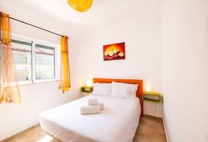 Una habitación de hotel con una cama, cortinas blancas, un cuadro y una mesita de noche con lámpara.