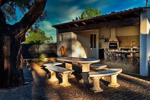 Un patio al aire libre con mesas y bancos de piedra, una pared de piedra y un edificio rústico con chimenea y asientos exteriores.