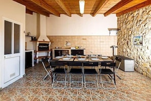 Un comedor con suelo de baldosas de piedra, una mesa redonda con sillas, una cocina con pared de piedra y un techo con vigas de madera.