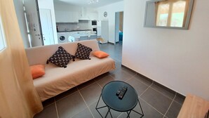 Ein Wohnzimmer mit einer Couch, zwei gemusterten Kissen, einem orangefarbenen Kissen, einem kleinen runden Tisch mit einer Fernbedienung und Blick in einen Küchenbereich mit einer Waschmaschine und einem Esstisch.