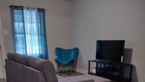 Uma sala de estar com um sofá modular cinza, uma poltrona azul, uma televisão sobre um móvel e uma janela com cortinas azuis.