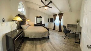 Quarto com cama grande, escrivaninha, cadeira, ventoinha no teto e janela com cortinas.