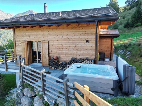 A wooden cabin with a hot tub, a wooden deck, and a mountain backdrop.
