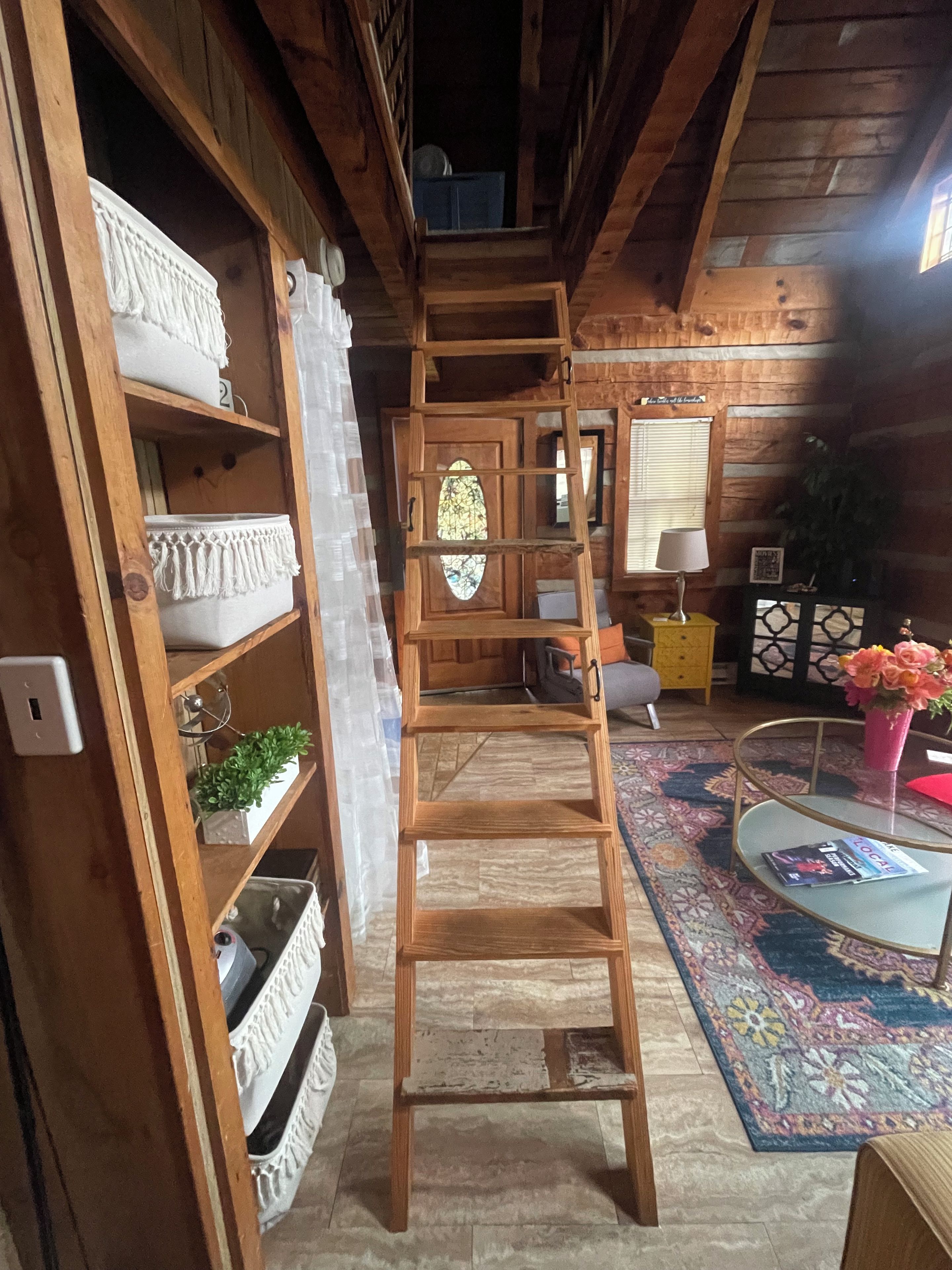 Un interior de madera con una escalera que lleva a un altillo, estantes con objetos decorativos, un espejo y una alfombra estampada.