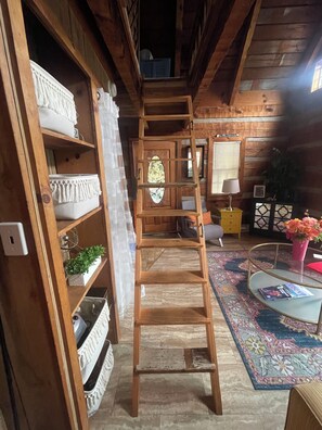 A wooden interior with a ladder leading to a loft, shelves with decorative items, a mirror, and a patterned rug.