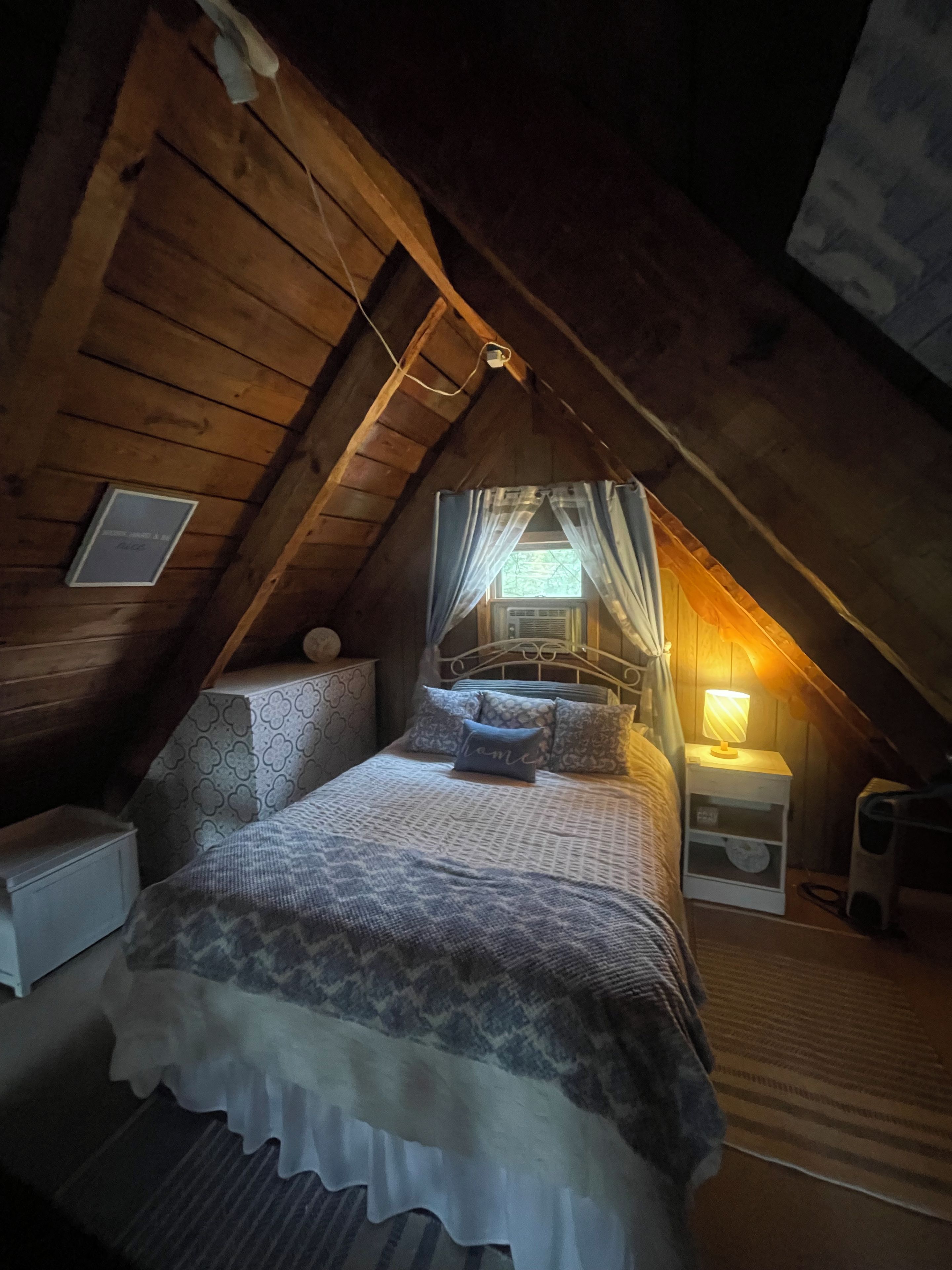A cozy attic bedroom with a sloped ceiling, a bed with patterned bedding, a nightstand with a lamp, and a window with sheer curtains.