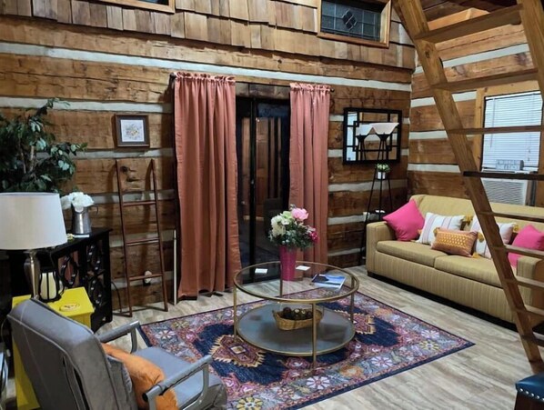 A cozy log cabin interior with a sofa, armchair, and a round coffee table with a vase of flowers. - Log Cabin on Main St. In the ❤️ of Winston Salem. Sleeps 4 comfortably.  (Winston-Salem)
