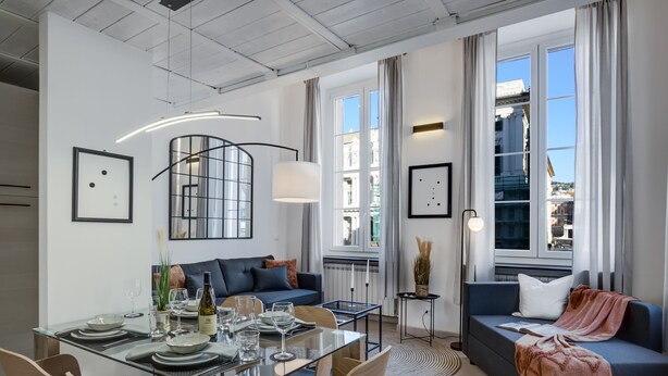 A modern living room with a glass dining table, a sofa, and a view of a cityscape through large windows.