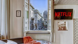 A hotel room with a bed, a window with a view of a city street, a wall-mounted TV with the Netflix logo, and a framed picture on the wall.