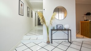 Un pasillo con suelo de baldosas, cuadros en las paredes, un espejo redondo y una pequeña mesa con un florero y objetos decorativos.