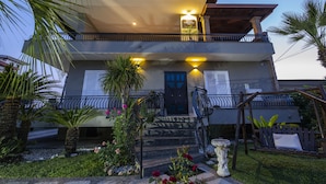 Un edificio de dos pisos con un balcón, una escalera y un jardín bien cuidado con palmeras y flores.