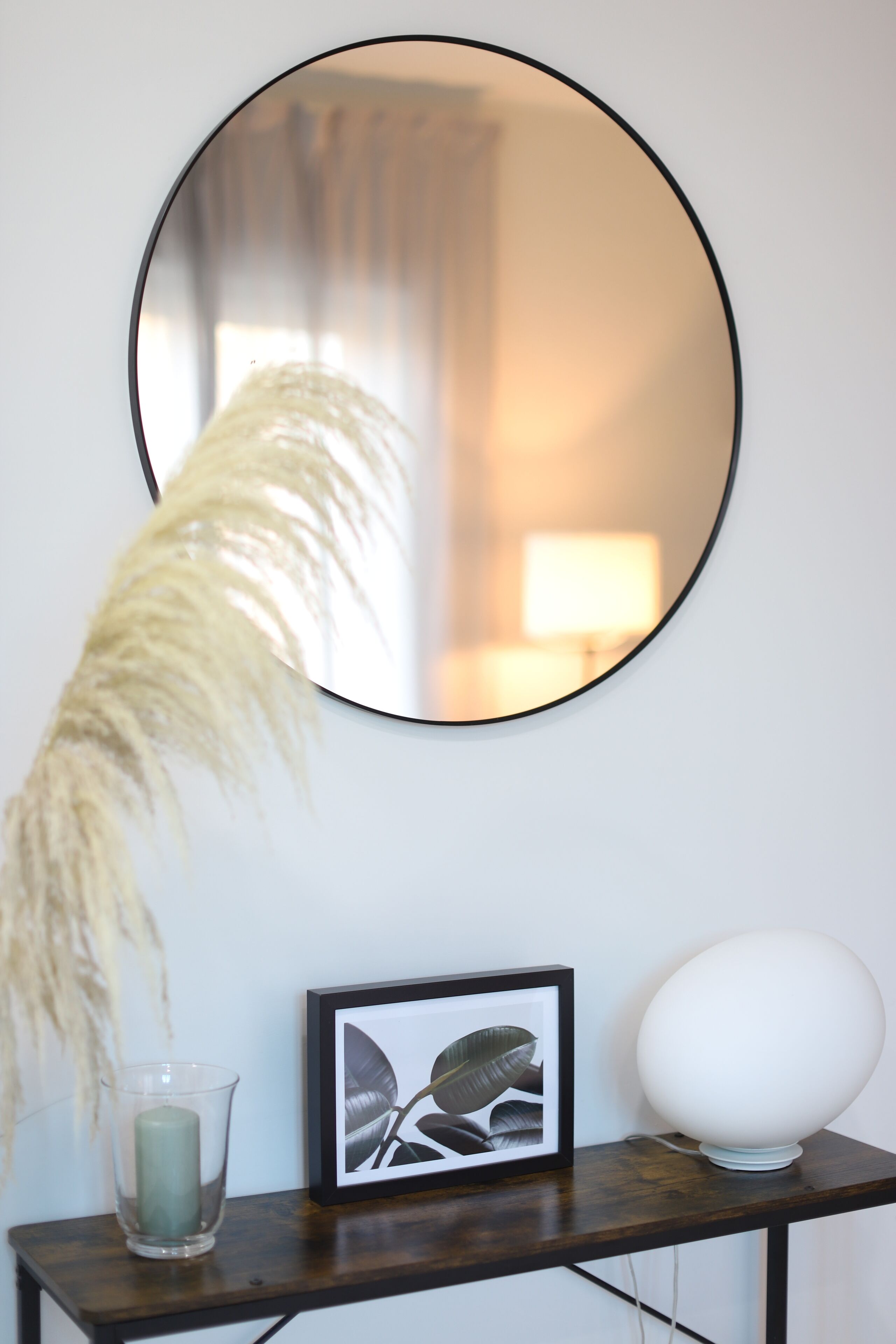 Un miroir rond accroché au mur, un tableau encadré, une lampe blanche et un vase en verre contenant une bougie, le tout posé sur une table en bois.