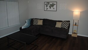 Een woonkamer met een donkerbruine hoekbank, een klein zwart salontafeltje en een lamp op een houten standaard.