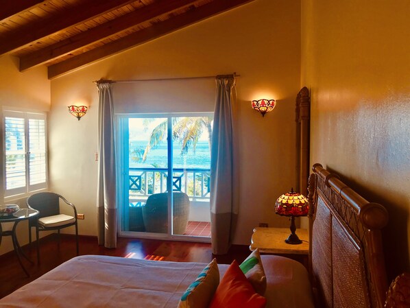 A hotel room with a bed, a chair, a table, a lamp, and a view of the ocean through a sliding door. - Sugar Mill hotel (West End)