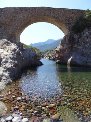 Eine steinerne Brücke spannt sich über einen klaren Fluss mit Kiesbett und Bergen im Hintergrund.