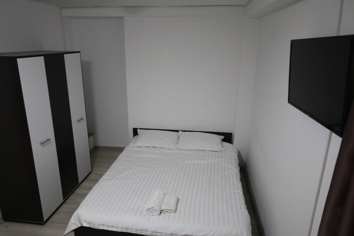 A small, neatly arranged bedroom with a bed, a wardrobe, and a flat-screen TV mounted on the wall.