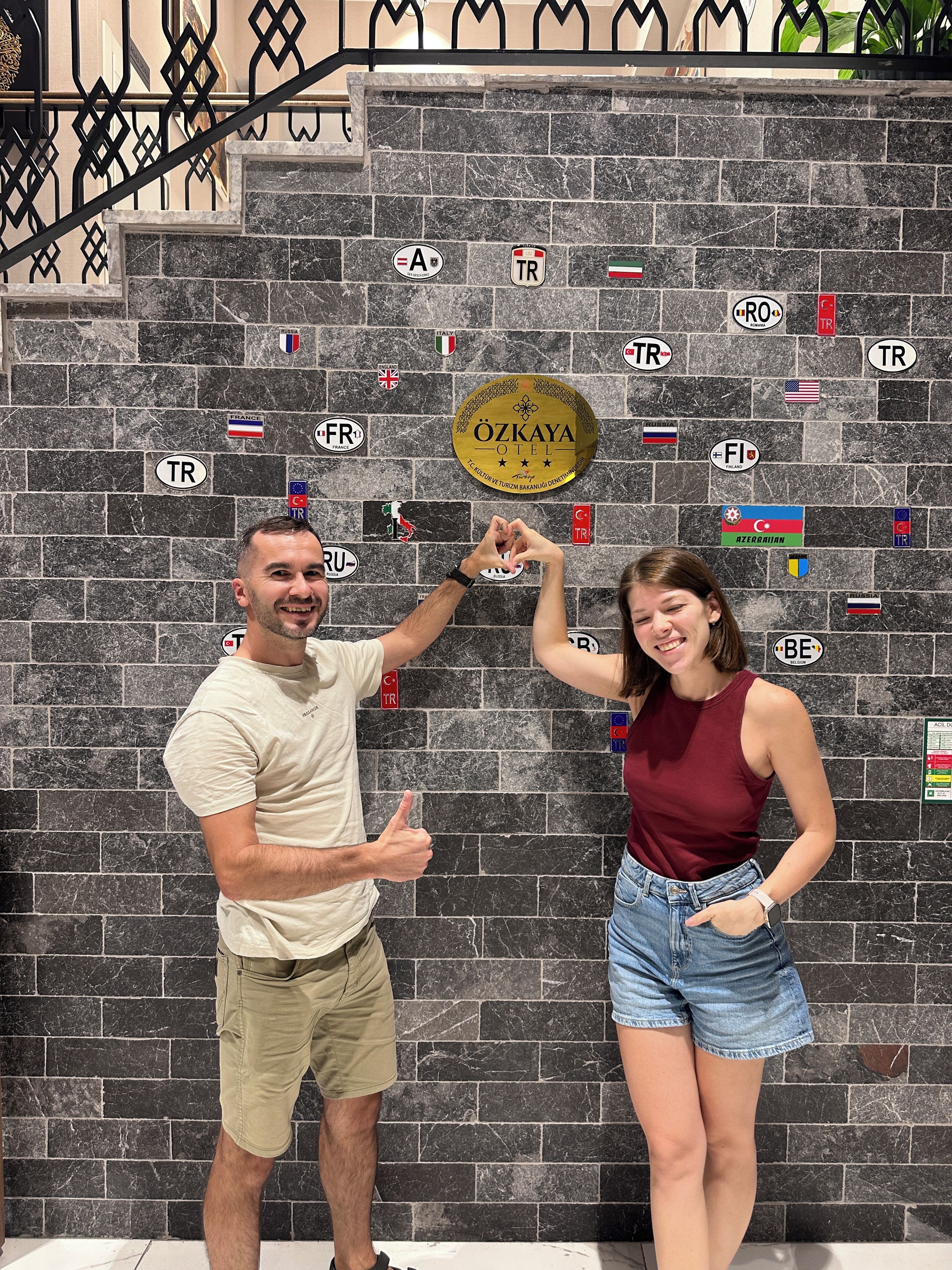 A man and a woman posing in front of a wall with various national flags and a sign that reads 'ÖZKAYA HOTEL'.