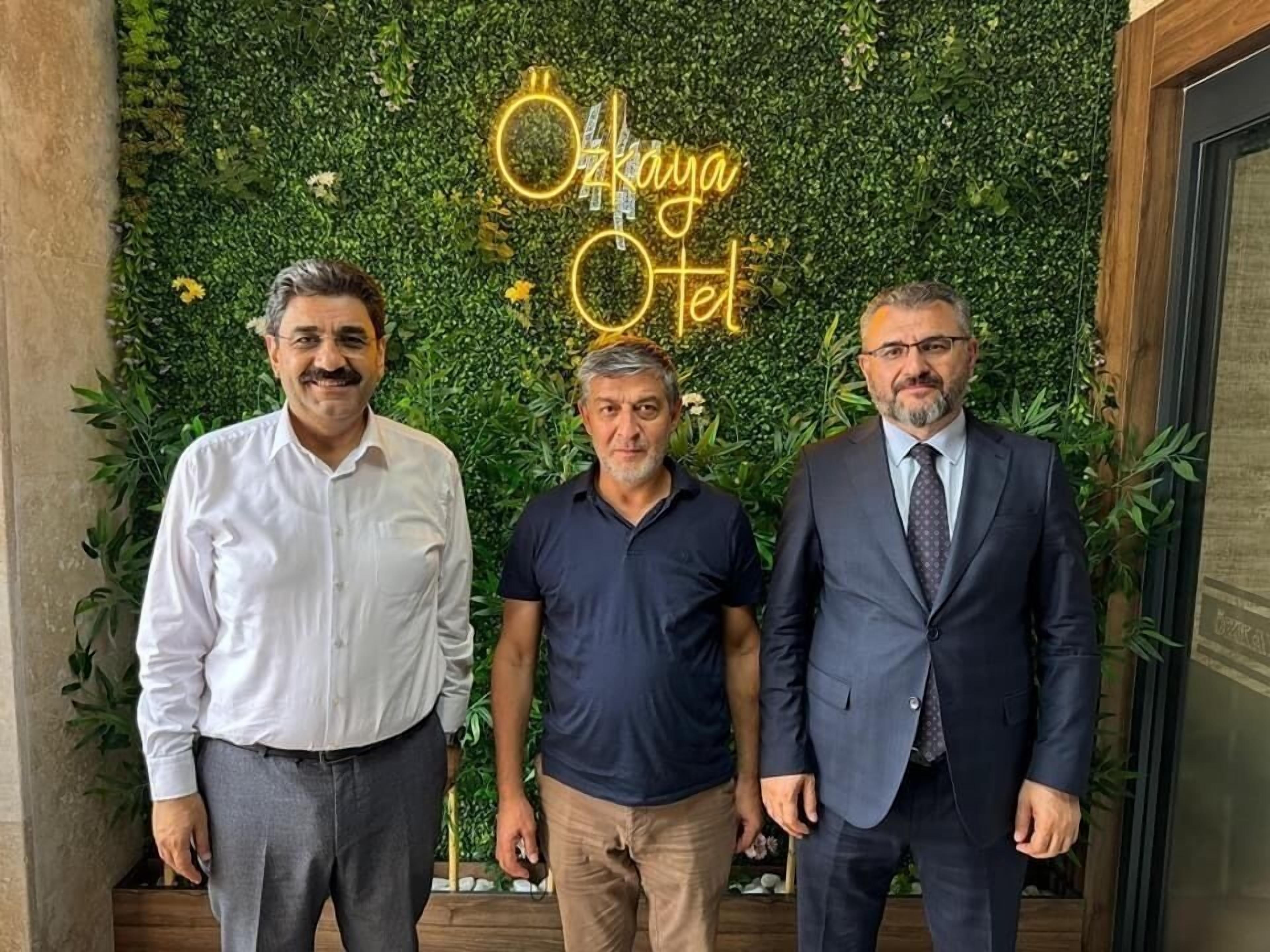 Tres hombres de pie frente a una pared con un letrero de neón que dice 'Okkaya Hotel'.