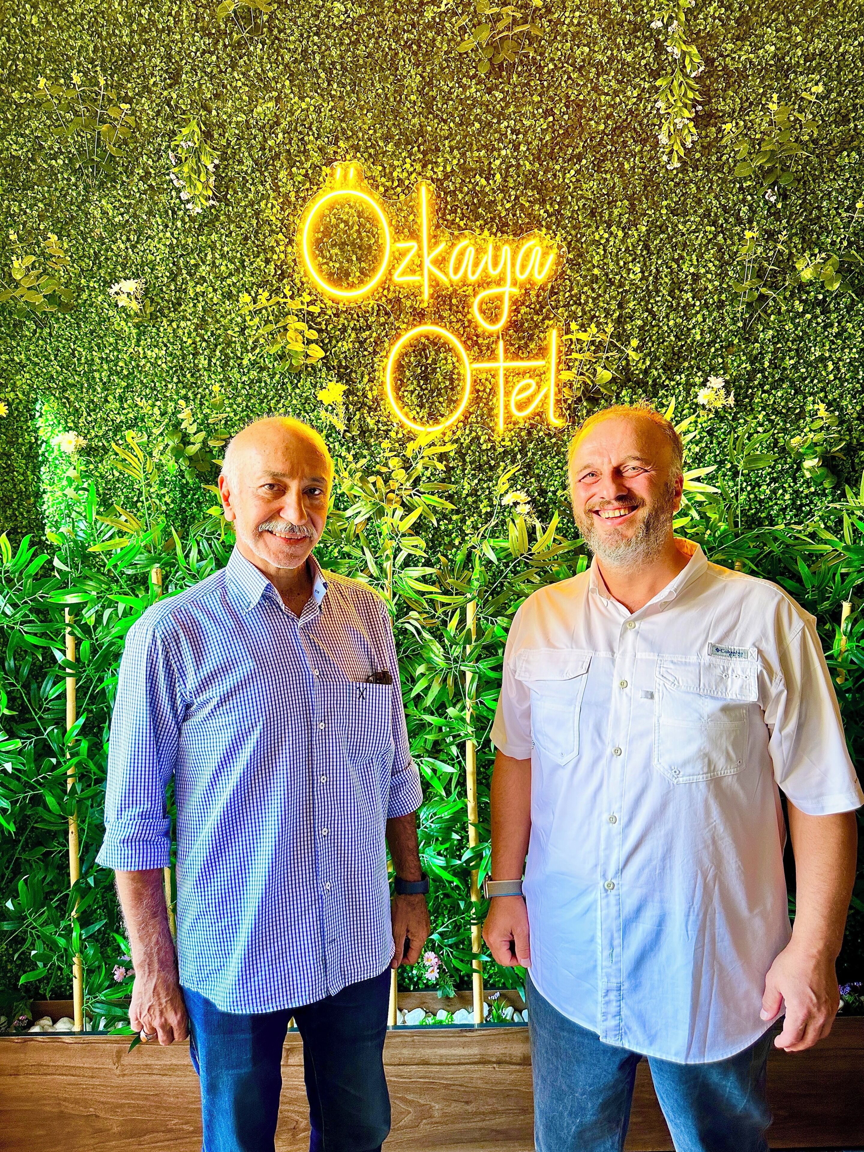 Two men standing in front of a lush green wall with a neon sign that reads "Ozkay Hotel".
