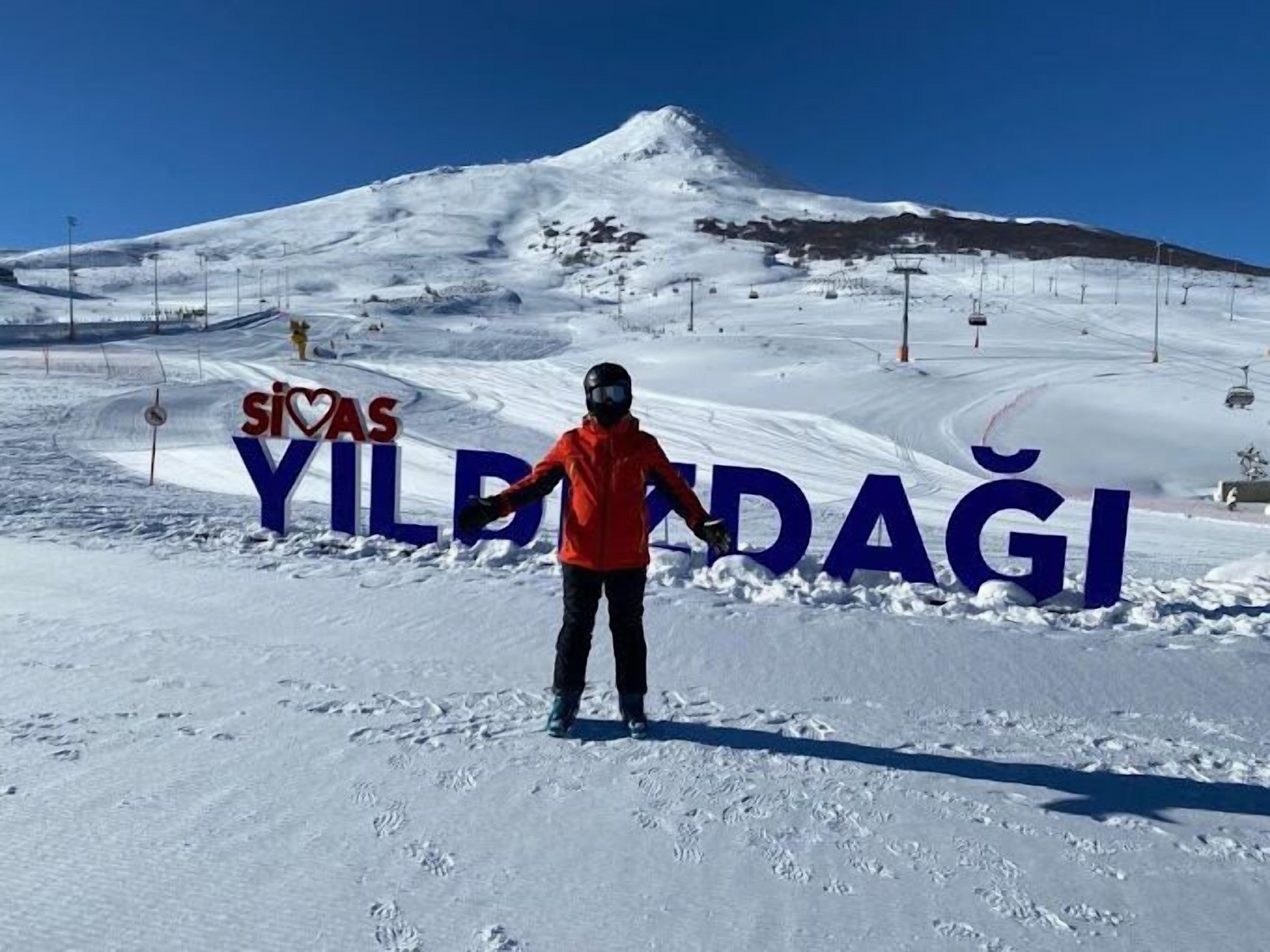 A person in a red jacket standing in front of a sign that reads "Sivas Yildagi".